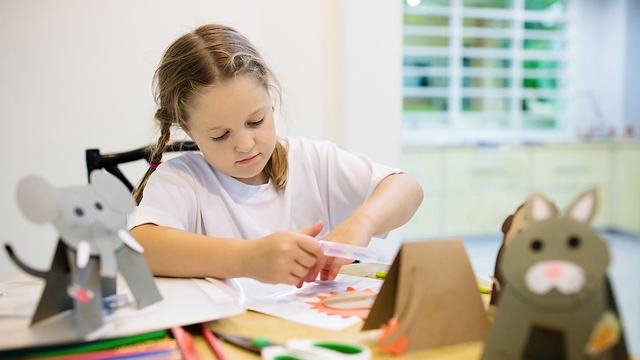 A small girl is completing a craft on a table covered in craft supplies, such as paper and scissors.