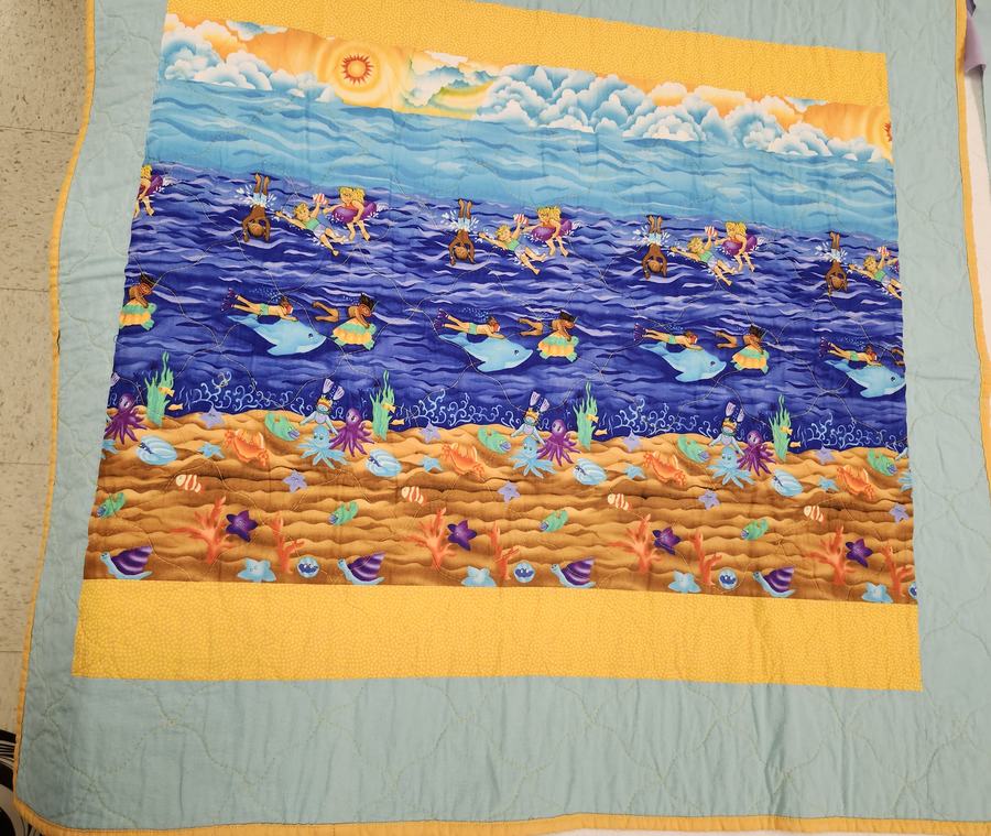 A baby quilt done in a blue and yellow color palette showing little kids swimming with dolphins.