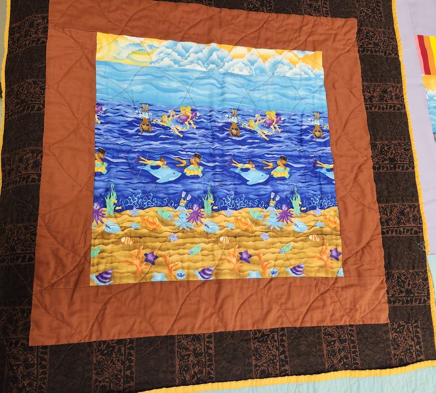 A baby NICO quilt done in a brown and gold color palette showing little kids swimming in the ocean with star fish and dolphins.