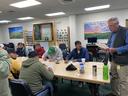 An older man reads papers to a group of men sitting at a table and writing in a classroom.