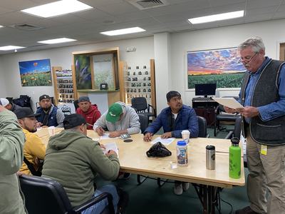 An older man reads papers to a group of men sitting at a table and writing in a classroom.
