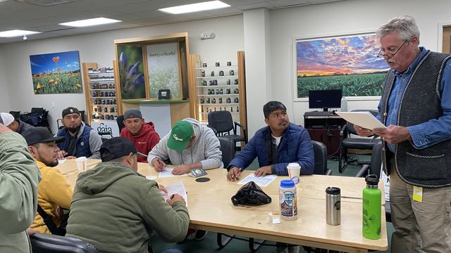 An older man reads papers to a group of men sitting at a table and writing in a classroom.