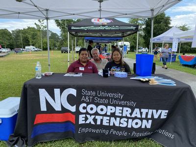 Three people sit behind an NC Cooperative Extension table at an outdoor community event.