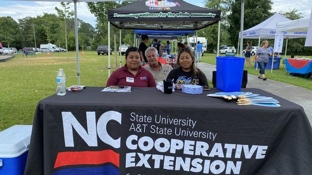 Three people sit behind an NC Cooperative Extension table at an outdoor community event.
