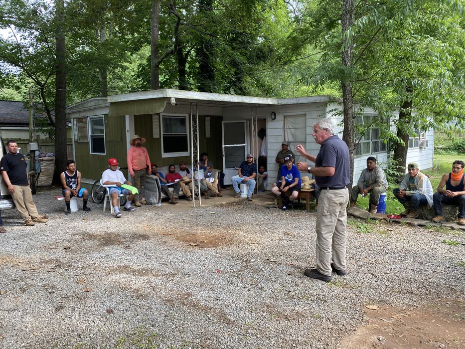 An older man speaks to a large group of men gathered outside a home in a wooded area.