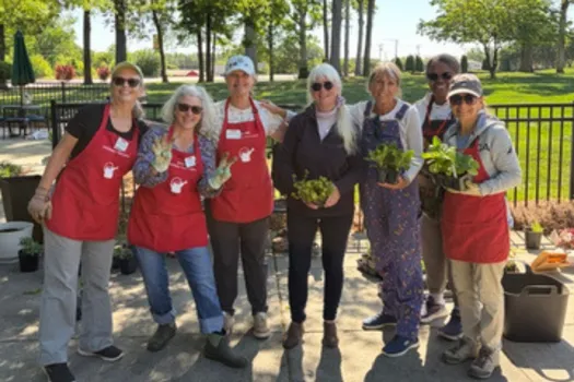 Six women stand together outdoors on a sunny day at the Jesse Helms Nursing Center, while holding plants and gardening supplies. Trees, a black fence, and patio seating are visible in the background during a community gardening project.