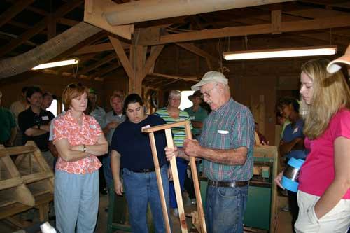 Older man demonstrating a wooden chair frame to a group inside a workshop