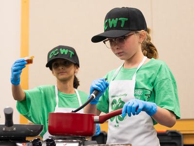 4-H members in matching green shirts and black caps participate in the Dinah Gore 4-H Healthy Food Challenge; one stirs a red saucepan with a spatula while another holds a small container. Both wear white aprons and blue gloves.