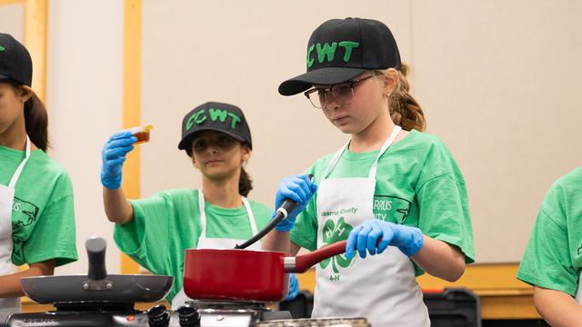 4-H members in matching green shirts and black caps participate in the Dinah Gore 4-H Healthy Food Challenge; one stirs a red saucepan with a spatula while another holds a small container. Both wear white aprons and blue gloves.