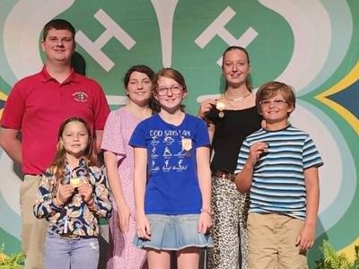 A group of six young individuals pose together on a stage in front of a large 4-H emblem backdrop, with several of them holding up medals.