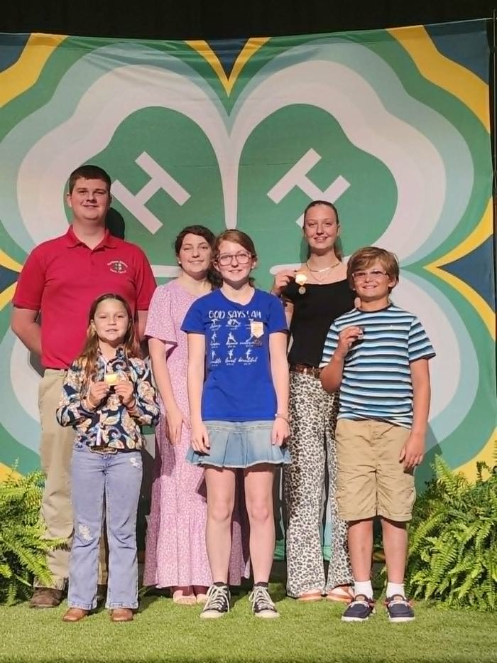 A group of six young individuals pose together on a stage in front of a large 4-H emblem backdrop, with several of them holding up medals.