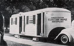A 1937 trailer hitiched to the back of a car. The trailer is marked with the words Electrification Electric Home equipment.