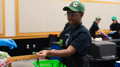 Young 4-H member in a green cap and glasses, wearing a black 4-H shirt and apron, holds a green shopping basket and uses a coin to buy items for their team at the Dinah Gore 4-H Healthy Food Challenge.