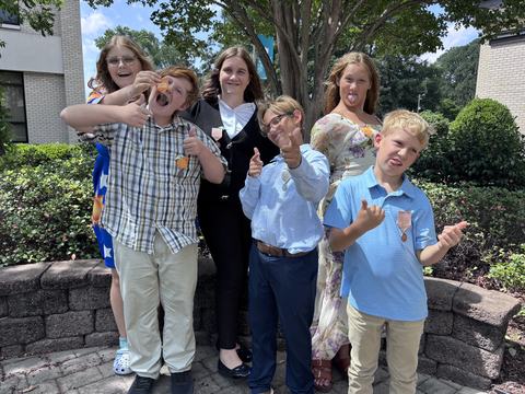 Six children pose outdoors in front of a tree and building, dressed in summer/formal clothes. They make playful faces and gestures, some wearing ribbons/medals.