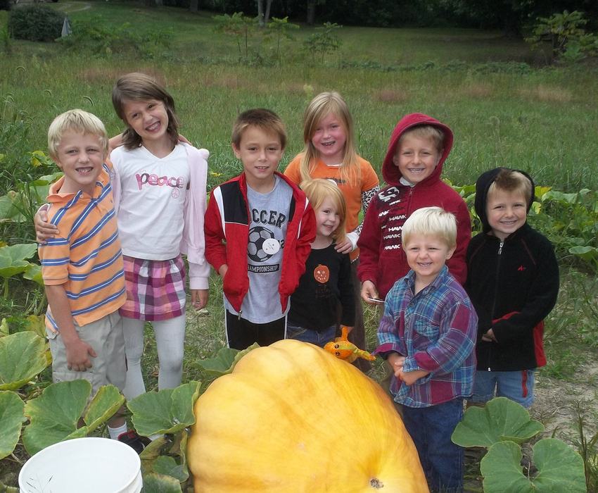 A group of youth standing behind a giant pumpkin in a field.