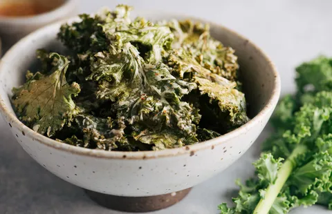 Image of a ceramic bowl full of kale chips topped with nutritional yeast, and to the right of the bowl, fresh green kale.