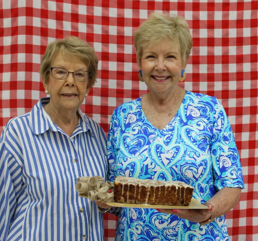 Two women stand in front of a red gingham background and smile while holding a apple dish