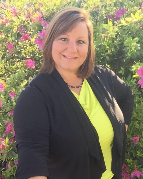 Dr. Kim Ingold posing with hands on hips in front of green bushes dotted with pink flowers.
