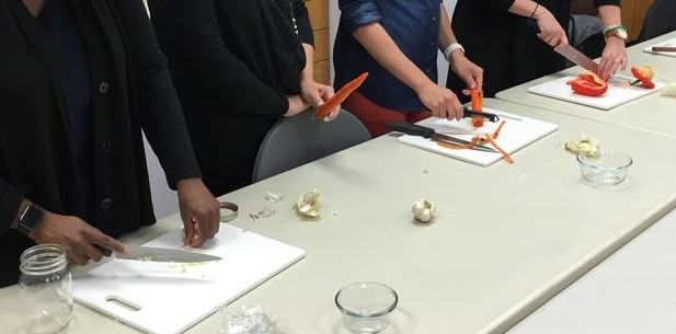 Five participants stand in a line behind a long table with a chopping board, a vegetable and a knife in hand to demonstrate their kife cutting skills.