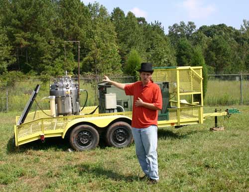 Man in red shirt and black hat gesturing toward yellow trailer with metal equipment