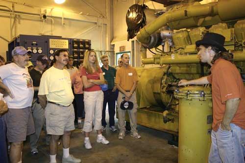 Man in hat explains large industrial machine to a group of standing visitors