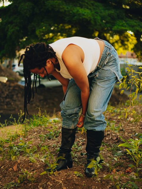 A young person with long braids leans over to tuck their pant legs into tall boots for gardening.