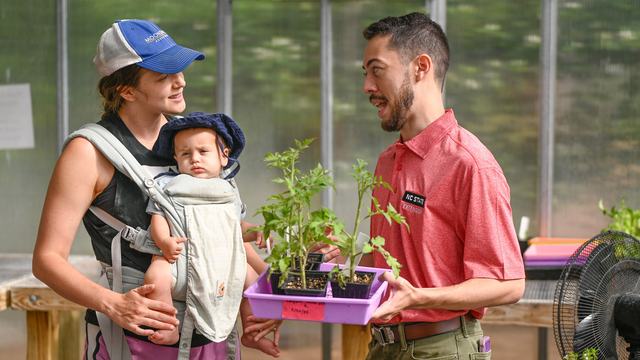 An NC State Extension agent helps a young mother with gardening seedlings while she holds her baby in a greenhouse.