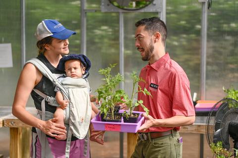 An NC State Extension agent helps a young mother with gardening seedlings while she holds her baby in a greenhouse.