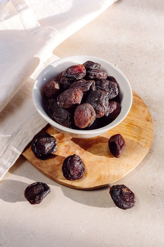 Dried prunes in a white bowl, set on a wooden board with natural lighting.