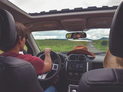 Male driving down long stretch of road with mountains in the distance and a cowboy hat on the dashboard of the car