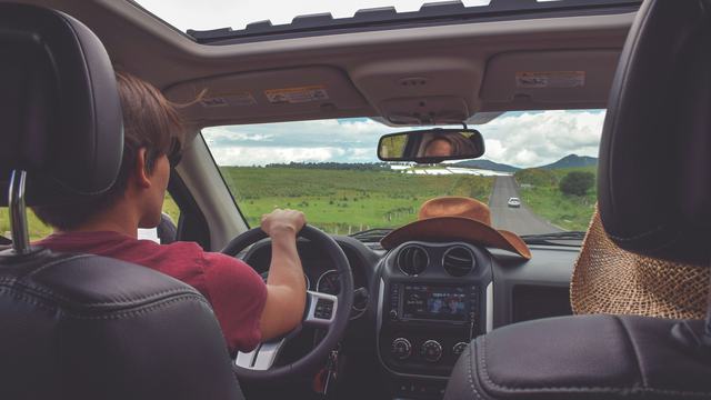 Male driving down long stretch of road with mountains in the distance and a cowboy hat on the dashboard of the car