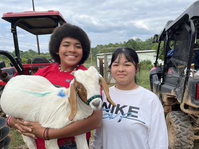 Two teenage girls stand next to each other smiling. One girl is holding a baby goat.