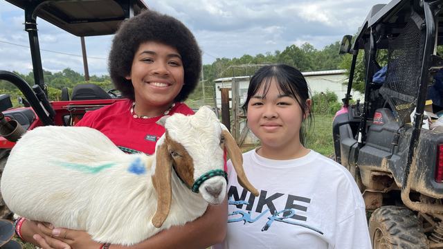 Two teenage girls stand next to each other smiling. One girl is holding a baby goat.