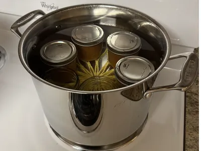 Boiling water bath canning of four jars of food and two empty jars in small pot on top of a yellow trivet.