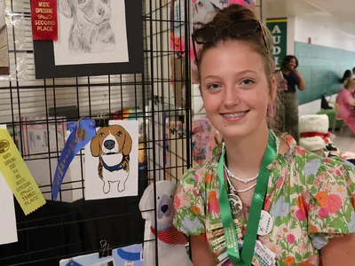 A 4-H member poses beside her award-winning artwork for the 4-H Dog Artistic Expression Contest displayed at the 2026 North Carolina 4-H State Presentation Finals.