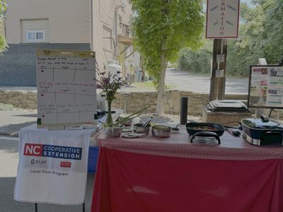picture of cooking demonstration at Marion Tailgate Market with red table cloth with checkered red top table cloth.  Bowls of foods on table with electric skillet and NC Cooperative Extension Local Food sign.