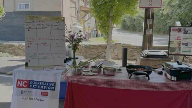 picture of cooking demonstration at Marion Tailgate Market with red table cloth with checkered red top table cloth.  Bowls of foods on table with electric skillet and NC Cooperative Extension Local Food sign.