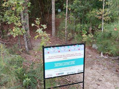 A sign at the entry of a wooded path encouraging people to read the story as they walk.
