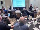 A presenter speaks to a group of Extension experts seated at round tables during a workshop, with attendees listening and viewing a presentation projected on a screen.