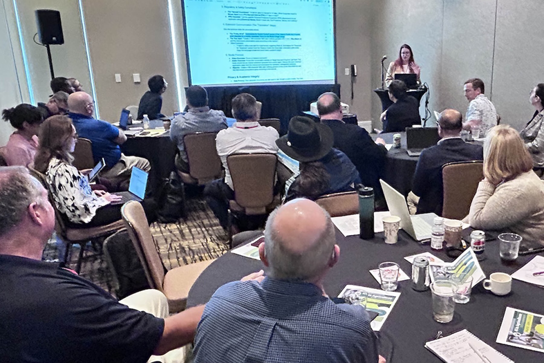 A presenter speaks to a group of Extension experts seated at round tables during a workshop, with attendees listening and viewing a presentation projected on a screen.