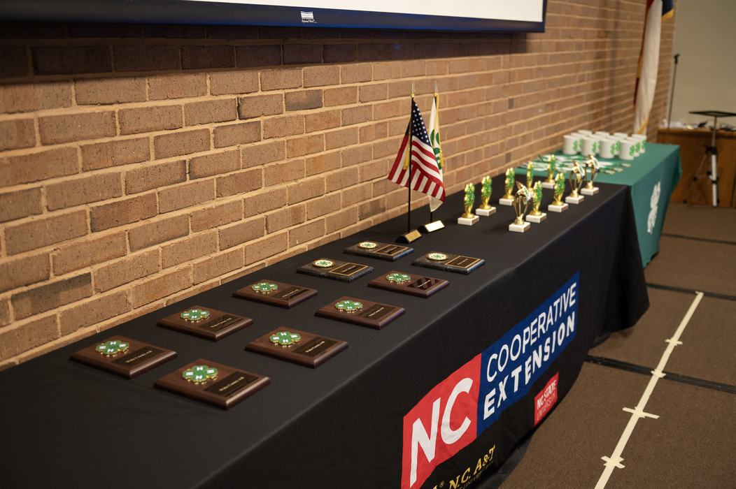 Table with NC Cooperative Extension Table Cloth and 4-H clovers and american flag and several plaques and statues for 4-h awards